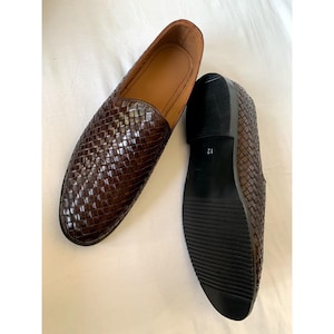 May include: A pair of dark brown woven leather loafers with a slip-on design. One shoe is positioned to show the textured upper, while the other is flipped to reveal the black sole with a visible size 12 marking on the heel area.