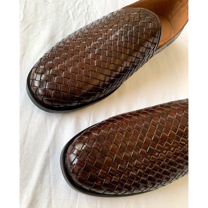 May include: A pair of dark brown leather loafers featuring a woven basket-weave pattern across the upper surface. The shoes have a rounded toe design, a low-profile silhouette, and a smooth tan interior lining visible at the heel opening.