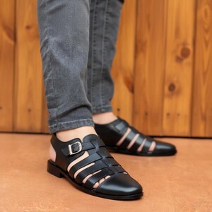 May include: A pair of black leather fisherman sandals featuring a closed toe, multiple wide straps across the upper, and an adjustable ankle strap with a metallic buckle. The sandals have a low heel and a smooth finish, providing a structured design.