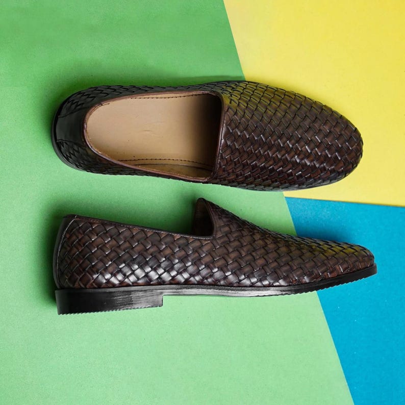 Pu&ograve; includere: Un paio di mocassini slip-on marrone scuro con una texture in pelle intrecciata sulla tomaia. Le scarpe presentano una silhouette a profilo basso con una fodera interna color beige liscia e una suola nera solida, viste dall'alto e di lato.