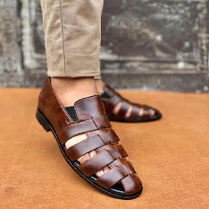 K&ouml;nnte beinhalten: Ein Paar braune Ledersandalen im Huarache-Stil mit geflochtenem Riemendesign auf der Oberseite, einer geschlossenen Ferse und einer schwarzen Sohle. Die Sandalen werden zu einer hellbeigen Hose getragen und zeigen die komplexe Flechtstruktur.