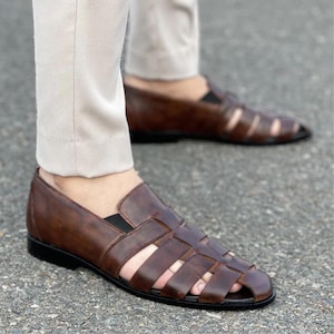 K&ouml;nnte beinhalten: Ein Paar dunkelbraune Fischer-Sandalen aus Leder mit geflochtenem Riemendesign, geschlossener Zehenpartie und niedrigem schwarzem Absatz. Die Sandalen werden zu einer hellen Hose getragen und zeigen einen Schlupf-Stil mit breiten Lederriemen.
