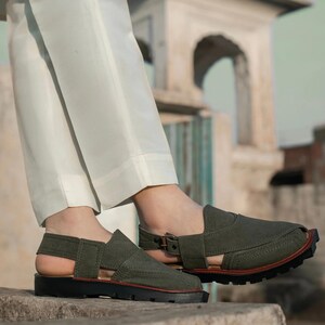 May include: A pair of olive green leather sandals with a buckle fastening and an open toe design. The sandals have a thick rubber sole and are being worn by someone with white trousers.