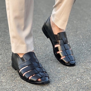 K&ouml;nnte beinhalten: Ein Paar schwarze Fischer-Sandalen aus Leder mit geflochtenem Riemendesign, geschlossener Ferse und abgerundeter Zehenpartie. Die Sandalen haben einen niedrigen Absatz und werden zu einer hellen Hose getragen, mit klassischem Cutout-Stil.