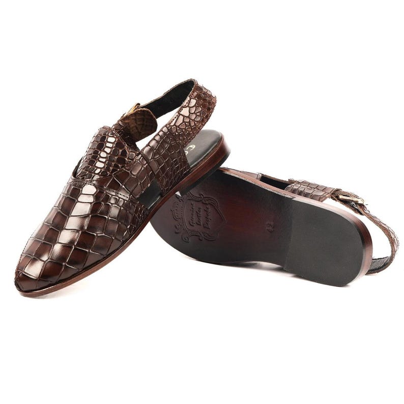 May include: A pair of dark brown slingback sandals featuring a glossy crocodile-embossed leather upper with a pointed toe. The sandals have a low heel, a buckled ankle strap, and a smooth dark brown sole with a visible embossed logo stamp.
