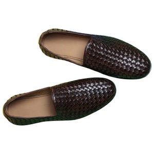 Pu&ograve; includere: Un paio di scarpe slip-on marrone scuro caratterizzate da una trama intrecciata sulla superficie superiore. Le scarpe presentano una punta arrotondata, un profilo basso e una fodera interna color cuoio chiaro, viste dall'alto su sfondo neutro.