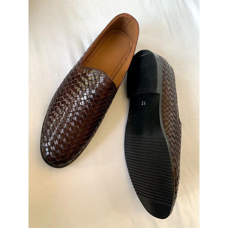Pu&ograve; includere: Un paio di mocassini slip-on in pelle intrecciata marrone scuro. Una scarpa &egrave; posizionata in modo da mostrare la fodera interna color cuoio, mentre l'altra &egrave; rivolta con la suola verso l'alto, rivelando una suola in gomma nera rigata con il numero 12.
