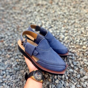 May include: A pair of blue leather sandals with a strap and buckle. The sandals have a brown sole and are on a gravel surface.