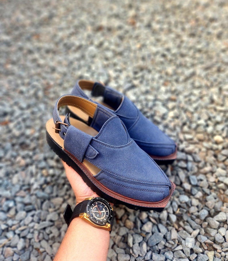 May include: A pair of blue leather sandals with a strap and buckle. The sandals have a brown sole and are on a gravel surface.