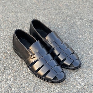 K&ouml;nnte beinhalten: Ein Paar schwarze Fischer-Sandalen aus Leder mit einem geflochtenen Riemendesign auf der Oberseite, einer geschlossenen Ferse und einer abgerundeten Zehenpartie. Diese Slipper haben eine glatte Oberfl&auml;che und ein flaches Profil mit breiten Riemen.