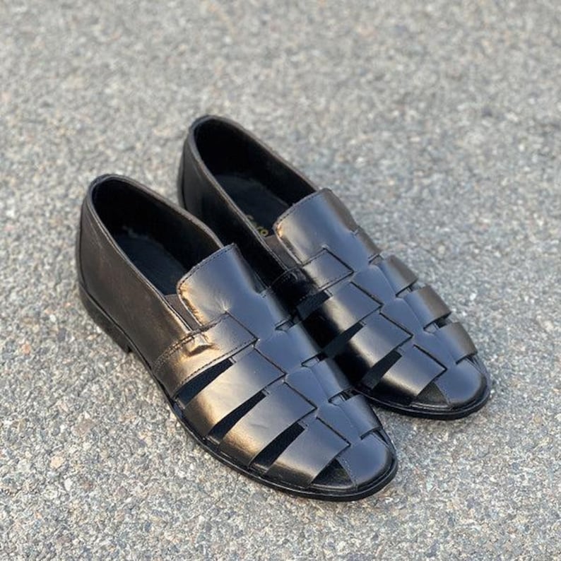 K&ouml;nnte beinhalten: Ein Paar schwarze Fischer-Sandalen aus Leder mit einem geflochtenen Riemendesign auf der Oberseite, einer geschlossenen Ferse und einer abgerundeten Zehenpartie. Diese Slipper haben eine glatte Oberfl&auml;che und ein flaches Profil mit breiten Riemen.