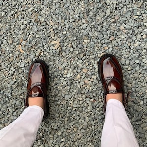 May include: A pair of brown leather shoes with a decorative strap and buckle. The shoes are worn by a person standing on a gravel surface.