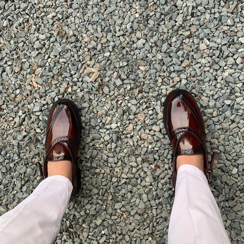 May include: A pair of brown leather shoes with a decorative strap and buckle. The shoes are worn by a person standing on a gravel surface.