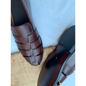 K&ouml;nnte beinhalten: Ein Paar dunkelbraune Leder-Sandalen im Huarache-Stil mit geflochtenem Obermaterial aus breiten Riemen und geschlossener Zehenpartie. Die Schuhe haben eine glatte Oberfl&auml;che, einen niedrigen Absatz und eine schwarze Sohle mit der Zahl 8.