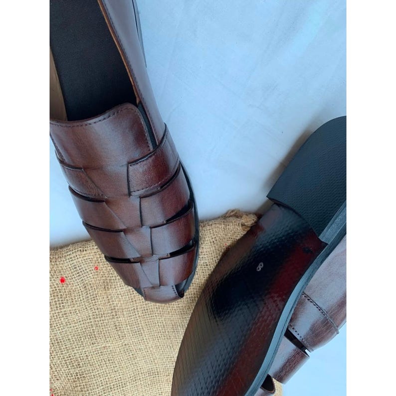 K&ouml;nnte beinhalten: Ein Paar dunkelbraune Leder-Sandalen im Huarache-Stil mit geflochtenem Obermaterial aus breiten Riemen und geschlossener Zehenpartie. Die Schuhe haben eine glatte Oberfl&auml;che, einen niedrigen Absatz und eine schwarze Sohle mit der Zahl 8.