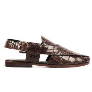 May include: A dark brown leather sandal with a crocodile-embossed texture. The sandal features a closed-toe design with side cutouts, a flat sole, and an adjustable ankle strap secured by a metallic buckle on the outer side of the footwear.