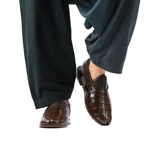 May include: A pair of dark brown leather sandals featuring a glossy, embossed crocodile skin texture. The footwear has a closed-toe design with woven straps, a slingback heel strap, and a flat sole, worn with loose-fitting dark charcoal trousers.