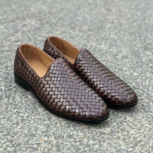 Pu&ograve; includere: Un paio di mocassini slip-on marrone scuro caratterizzati da una tomaia in pelle intrecciata. Le scarpe presentano un design a profilo basso con una fodera interna liscia e una suola sottile di colore scuro, con un motivo intrecciato uniforme.