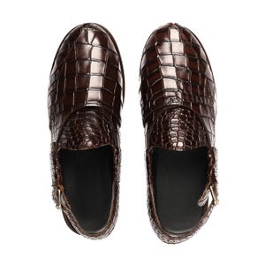 May include: A pair of dark brown slip-on shoes featuring a glossy crocodile-embossed texture. The shoes have a rounded toe, a solid black interior lining, and a small metallic buckle detail on the side of each shoe, shown from a top-down view.