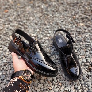 May include: A pair of black leather slip-on shoes with a buckle strap and a brogue design. The shoes have a glossy finish and a thick sole.