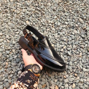 May include: Black leather slip-on sandal with a buckle closure and a brogue design. The sandal has a thick black sole.