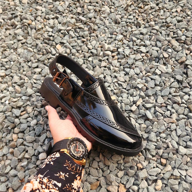 May include: Black leather slip-on sandal with a buckle closure and a brogue design. The sandal has a thick black sole.