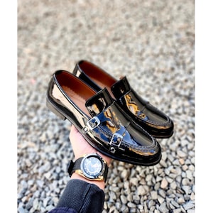 Pode incluir: Um par de sapatos de mocassim pretos de verniz com fivelas duplas. Os mocassins têm uma ponta pontiaguda e um salto baixo.