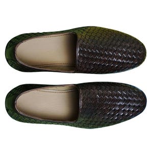 Pu&ograve; includere: Un paio di scarpe slip-on marrone scuro caratterizzate da una trama intrecciata sull'esterno. Le scarpe presentano una punta arrotondata e una fodera interna liscia color beige chiaro visibile dall'alto, che mette in risalto il motivo.