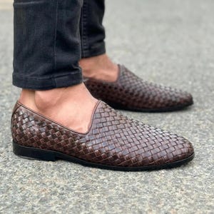 Pu&ograve; includere: Un paio di mocassini in pelle intrecciata marrone scuro con design slip-on, profilo basso e suola nera solida. La parte superiore della scarpa &egrave; realizzata con un motivo a intreccio stretto e complesso che copre l'intera superficie esterna.