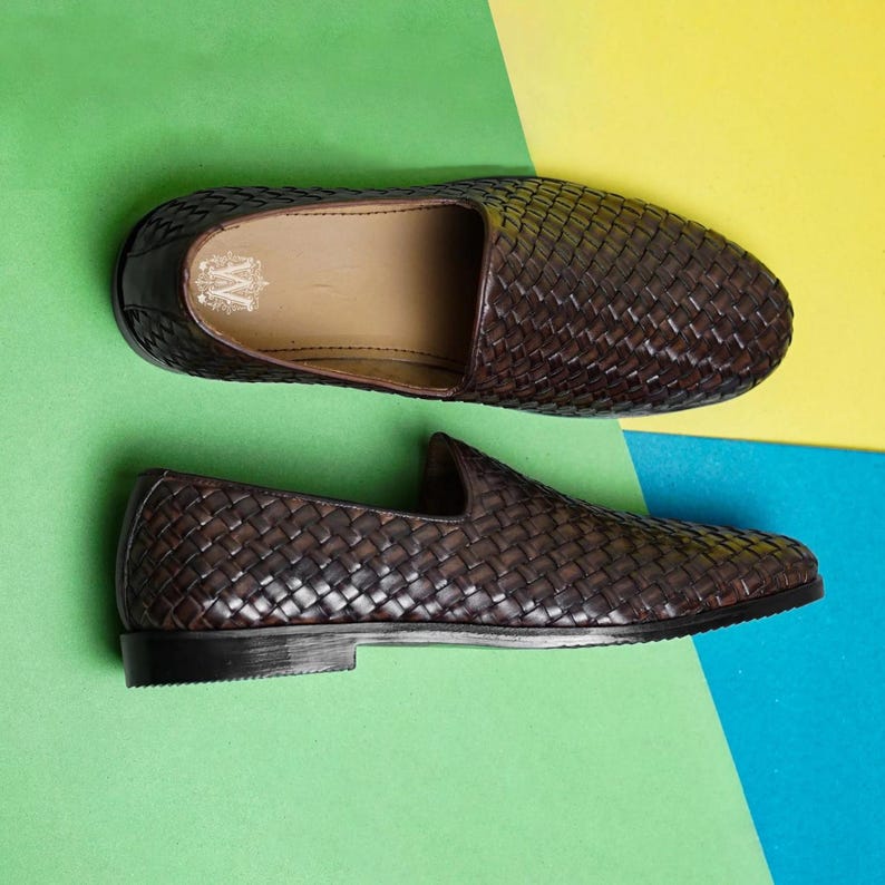May include: A pair of dark brown woven leather loafers featuring a slip-on design with a low heel and a rounded toe. The interior of the shoe displays a white logo with the letter W printed on the tan insole, showcasing a detailed basket-weave texture.