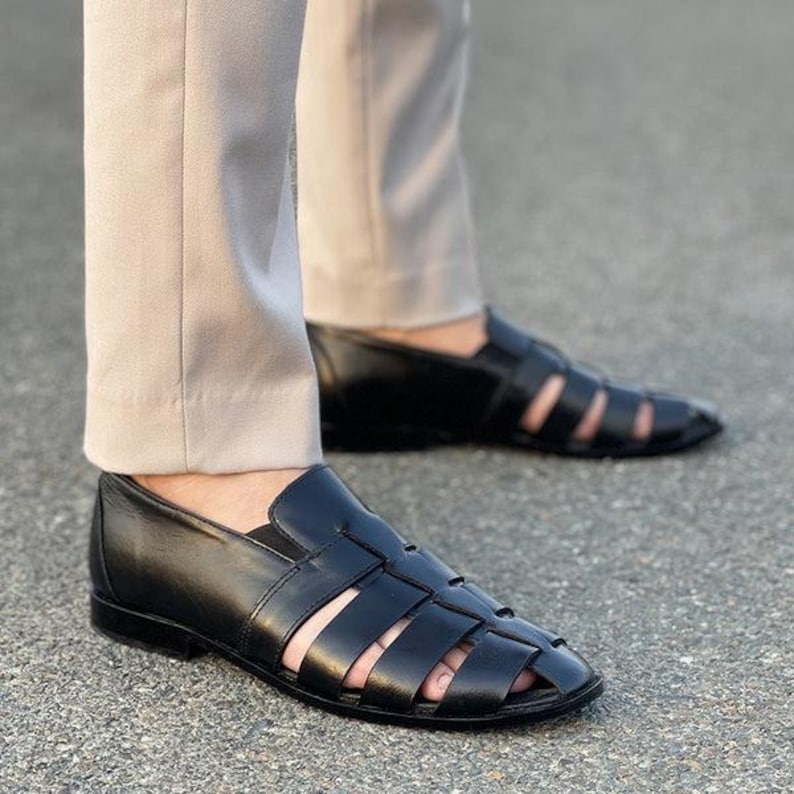 K&ouml;nnte beinhalten: Ein Paar schwarze Fischer-Sandalen aus Leder mit geschlossener Zehenpartie, einer Reihe breiter geflochtener Riemen am Obermaterial und einem niedrigen Absatz. Die Sandalen werden zu einer hellbeigen Hose getragen und wirken klassisch.