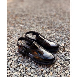 May include: A pair of black leather shoes with a pointed toe and a strap around the ankle. The shoes have a decorative pattern on the front and are sitting on a bed of small stones.