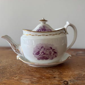 Puede incluir: Tetera de porcelana blanca con tapa y platillo a juego. La tetera presenta un diseño de querubines morados y ribetes dorados. El pico y el asa tienen detalles dorados decorativos. La tetera está sobre una superficie de madera.