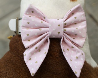 Op de afbeelding: Een roze hondenstrik met een witte halsband. De strik is bedrukt met kleine witte en gouden hartjes. De strik is bevestigd aan een witte halsband met een zilveren gesp. De vacht van de hond is bruin en wit.