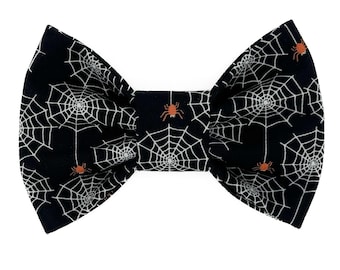 May include: A black bow tie featuring a white spider web design and small orange spiders. This bow tie is a Halloween-themed accessory.