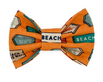 May include: Orange bow tie with a beach-themed design. The bow tie features directional signs with the word "BEACH" in white block letters. The signs point to the beach, with text such as "TO THE BEACH" and "EACH IS WAY".