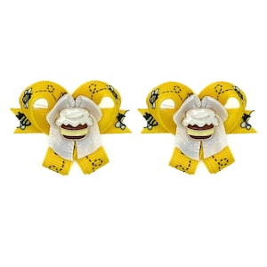 May include: Two yellow hair bows with a bee and honeycomb design. Each bow features a white center with a honey pot charm. The bows are made of ribbon and are set against a white background.