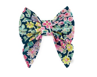 May include: A large, fabric bow with a floral pattern. The bow features a dark teal background with pink, yellow, and purple flowers and green leaves. The bow is tied in the center and has two long, flowing ends.