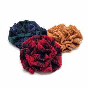 May include: Three fabric flower brooches in a variety of patterns. One is red and black plaid, another is blue and green plaid, and the third is a tan herringbone weave. The brooches are arranged on a white background.