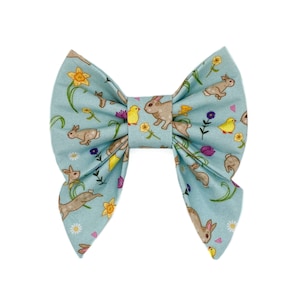 May include: A light blue fabric bow with a repeating pattern of brown bunnies, yellow chicks, and colorful flowers. The bow is tied in the center and has a classic bow shape, perfect for Easter or spring.
