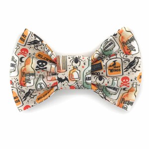 Può includere: Un papillon a tema Halloween con sfondo color crema e un motivo ripetuto di bottiglie di pozioni, teschi, ragni, pipistrelli e corvi. Le bottiglie sono etichettate con parole come "Veleno", "Arsenico" ed "Elisir".