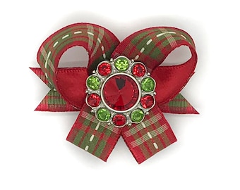 May include: A festive Christmas bow with a red satin ribbon and a plaid ribbon in red, green, and white. The bow features a central embellishment with a large red gem surrounded by smaller red and green gems.