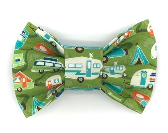 May include: A green bow tie with a repeating pattern of camping trailers, tents, and trees. The bow tie is made of fabric and has a classic bow shape. The colors include green, white, blue, and orange.