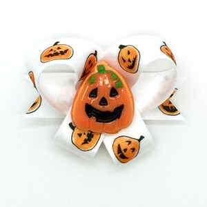 Puede incluir: Un lazo de satén blanco con calabazas naranjas estampadas en la cinta. Una calabaza naranja grande con una cara sonriente está centrada en el lazo. Accesorio temático de Halloween.