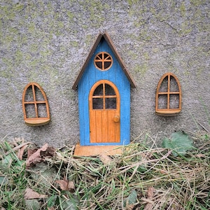 Può includere: Una casetta di legno blu in miniatura con una porta e finestre in legno marrone. La casetta è attaccata a un muro di cemento grigio con muschio verde.