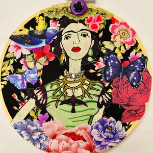 May include: A round embroidery hoop with a black background featuring a colourful floral design. The centre of the hoop features a portrait of Frida Kahlo with a green and black bird, butterflies, and flowers surrounding her.
