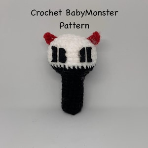 May include: A handmade crochet BabyMonster pattern in black, white, and red. The pattern features a white head with black details and red horns, atop a black handle. The text "Crochet BabyMonster Pattern" is above the crochet figure.