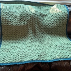 May include: A handmade, textured blanket in shades of green with a teal blue border. The blanket is made of soft yarn and has a woven pattern. The blanket is draped over a dark brown leather couch.