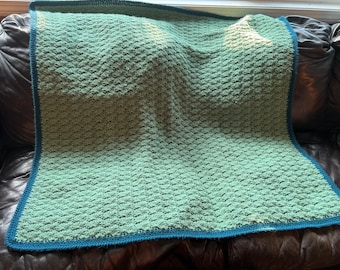 May include: A hand-knitted blanket in shades of green with a teal blue border. The blanket has a textured, woven pattern. The blanket is made of soft yarn and is draped over a dark brown leather sofa.