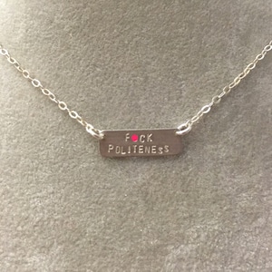 Peut inclure: Un collier en argent avec un pendentif rectangulaire qui dit "F*CK POLITENESS" en lettres noires avec un point rouge au-dessus du "O".
